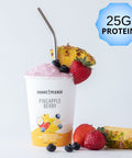 Premade pineapple berry protein smoothie in a white cup with a yellow lid labeled 25g protein, with fresh pineapple chunks, strawberries, blueberries, and banana slices around it.